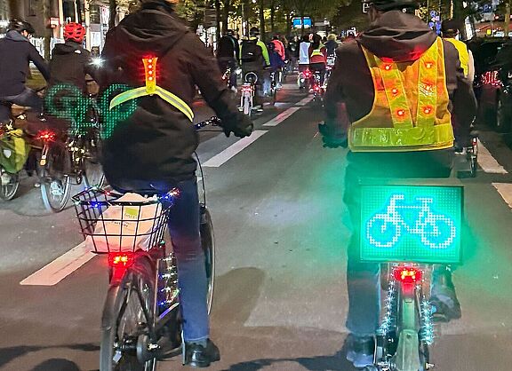ADFC-Lichterfahrt Berlin, 10. Oktober 2025: Mehrere Menschen fahren auf beleuchteten Fahrrädern durch eine Fußgängerzone. An den Fahrrädern leuchten rote und grüne LED-Lichter, ein Fahrrad trägt ein leuchtend grünes Display mit Fahrrad-Symbol. Die Radfahrenden tragen fluoreszierende Sicherheitswesten. Im Hintergrund sind weitere beleuchtete Fahrräder zu sehen.